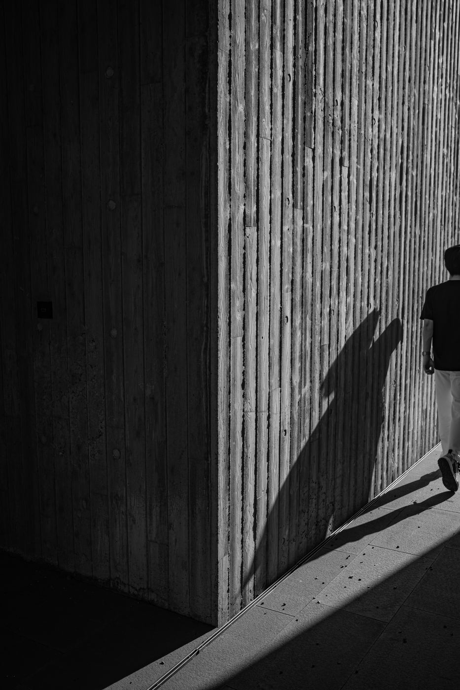 Person walking past a ribbed concrete wall in black and white, long shadow across the pavement